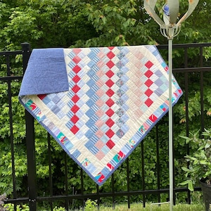 May include: A handmade quilt with a blue backing and a patchwork design of red, white, and blue squares. The quilt is draped over a black metal fence outdoors, showcasing its vibrant colors and intricate pattern.