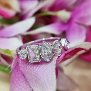 May include: A silver ring featuring five clear gemstones of varying shapes and sizes. The ring is displayed against a pink fabric backdrop.