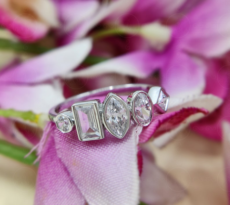 May include: A silver ring featuring five clear gemstones of varying shapes and sizes. The ring is displayed against a pink fabric backdrop.