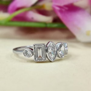 May include: A silver ring featuring five clear gemstones of varying shapes. The ring includes a round stone, a rectangular stone, an oval stone, and two smaller round stones.