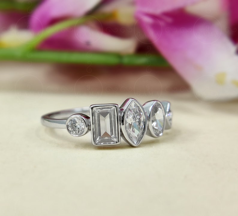 May include: A silver ring featuring five clear gemstones of varying shapes. The ring includes a round stone, a rectangular stone, an oval stone, and two smaller round stones.