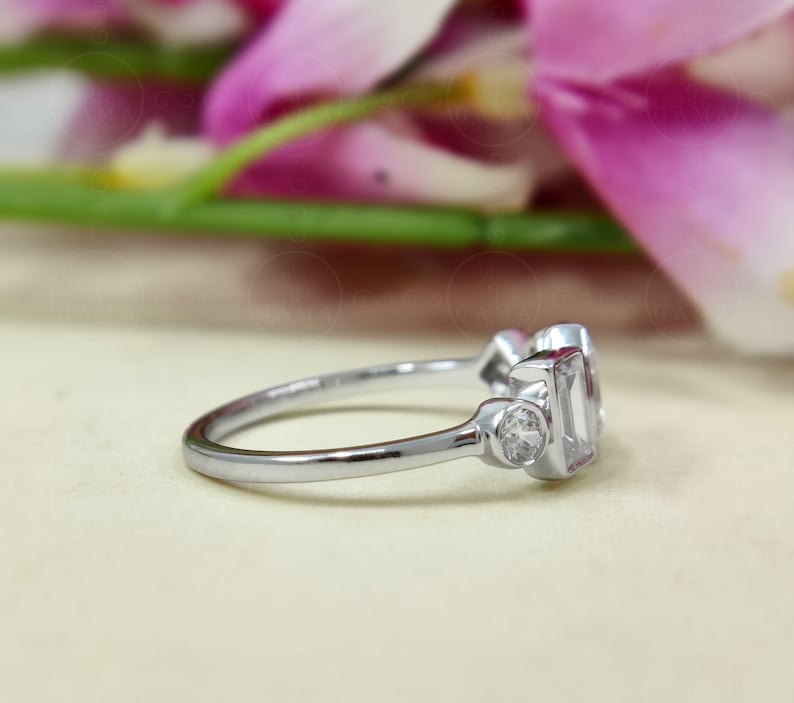 May include: A silver ring featuring three clear gemstones. The central stone is a rectangular emerald cut, while the two side stones are round brilliant cuts.