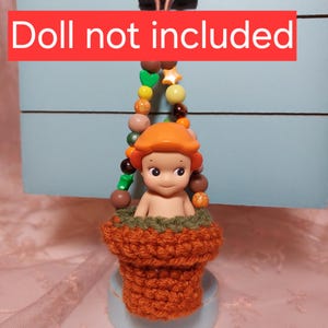 May include: A crocheted flower pot with a green and orange color scheme. The pot is hanging from a beaded chain with green, yellow, and orange beads. The text "Doll not included" is visible in the image.