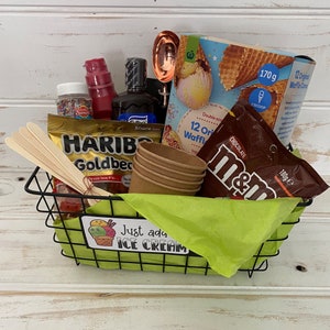 May include: A black wire basket filled with a variety of items for making ice cream sundaes. The items include waffle cones, chocolate candies, sprinkles, a small bottle of chocolate syrup, a red bottle of liquid, wooden spoons, and paper cups. The basket is sitting on a white wooden surface. The text "Just add ice cream" is printed on a green sticker on the front of the basket.