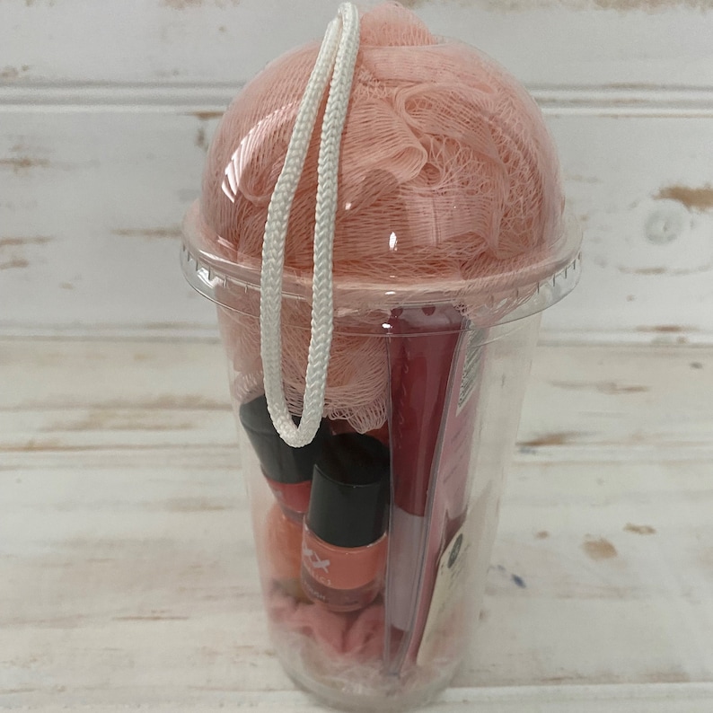 May include: A clear plastic cup filled with bath and beauty products. The cup contains a pink bath pouf, two bottles of nail polish, and a red and white striped item. The cup has a white drawstring.