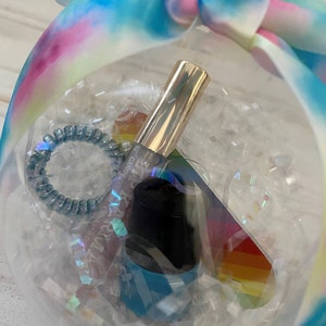 Puede incluir: Una esfera de pl&aacute;stico transparente llena de papel de seda blanco y una tarjeta de arco&iacute;ris. Dentro de la esfera hay una botella de esmalte de u&ntilde;as azul, un tubo de brillo de labios dorado y una liga para el cabello en espiral azul.