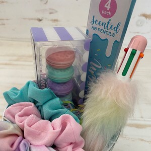 K&ouml;nnte beinhalten: Ein Geschenkkorb gef&uuml;llt mit pastellfarbenen Artikeln, darunter eine Packung mit 4 duftenden HB-Stiften, ein mehrfarbiger Stift mit einem flauschigen Regenbogen-Pompon, ein Set mit vier pastellfarbenen Macaron-f&ouml;rmigen Radiergummis und ein Set mit drei pastellfarbenen Haargummis.