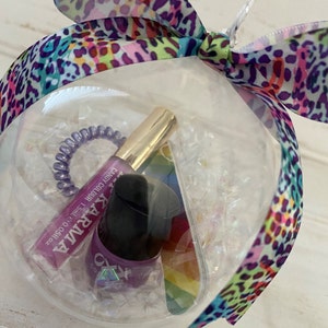 Puede incluir: Un adorno de pl&aacute;stico transparente lleno de peque&ntilde;os confeti iridiscentes, una liga para el cabello morada, una peque&ntilde;a botella de brillo labial rosa etiquetada como "KARMA CANDY COLOUR 1.5ml 0.05fl oz", y una peque&ntilde;a botella de esmalte de u&ntilde;as morado con una pegatina de arco&iacute;ris en el lateral.