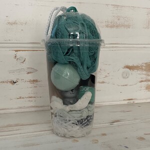 May include: A clear plastic cup filled with bath products, including a green bath pouf, a green bath bomb, and white bath confetti.
