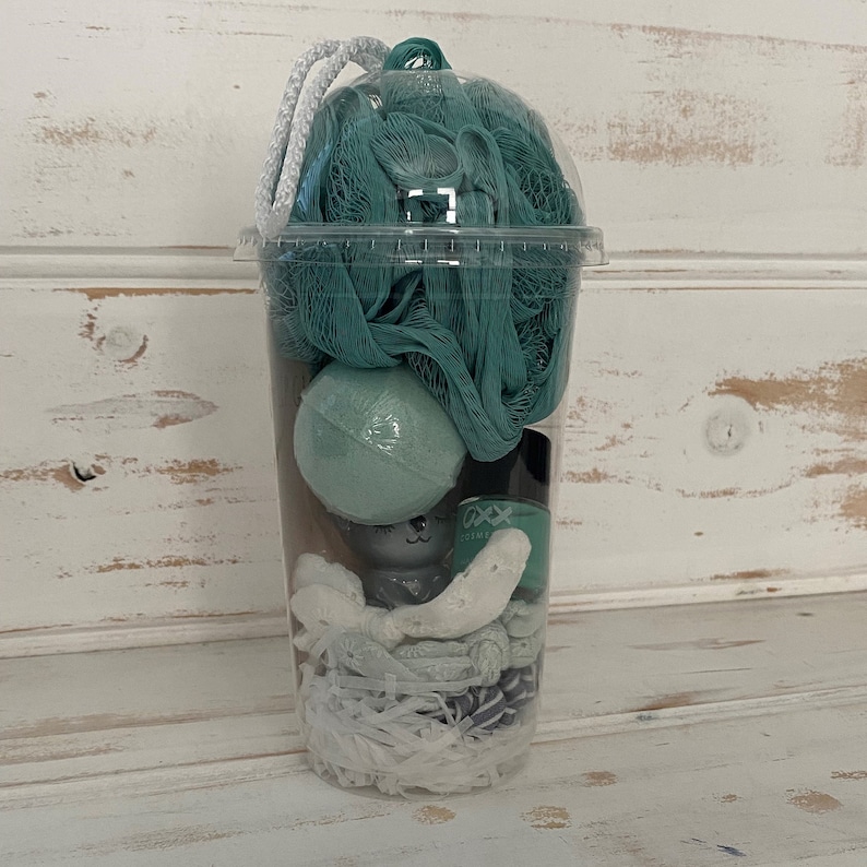 May include: A clear plastic cup filled with bath products, including a green bath pouf, a green bath bomb, and white bath confetti.