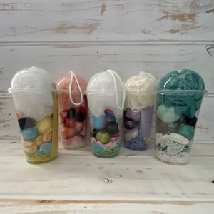 May include: Four clear plastic cups filled with various bath and beauty products. The cups are decorated with colorful ribbons and have lids. The products include bath bombs, bath salts, soap, and lotion.