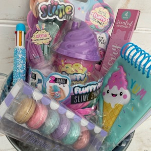 K&ouml;nnte beinhalten: Ein Geschenkkorb gef&uuml;llt mit verschiedenen Artikeln, darunter ein lila Schleimbeh&auml;lter, ein Notizbuch mit einem Comic-Eiswaffel, eine Packung Duftbleistifte und eine Packung Macaron-Schleim. Der Korb ist mit blauem und rosa Seidenpapier gef&uuml;llt.