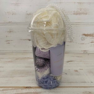 May include: A clear plastic cup filled with bath products. The cup contains a white bath puff, a purple bath bomb, a small bottle of purple liquid, a tube of hand cream labeled "Karma Hand Cream Coconut & Vanilla", and purple shredded paper.