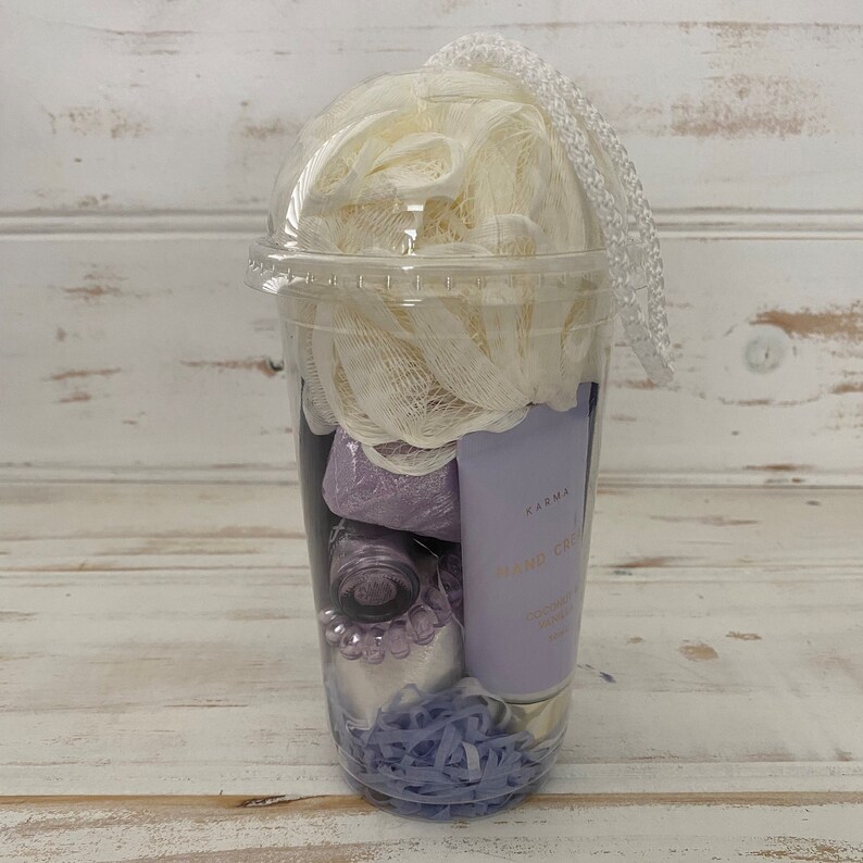 May include: A clear plastic cup filled with bath products. The cup contains a white bath puff, a purple bath bomb, a small bottle of purple liquid, a tube of hand cream labeled "Karma Hand Cream Coconut & Vanilla", and purple shredded paper.