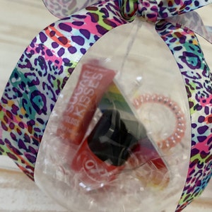 Puede incluir: Un adorno de pl&aacute;stico transparente lleno de un tubo de brillo de labios rojo con el texto "Sugar Kisses", una pinza para el pelo negra y una liga para el pelo rosa.