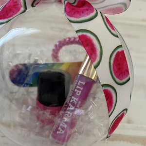 Puede incluir: Un adorno de pl&aacute;stico transparente lleno de papel de seda blanco y botellas de esmalte de u&ntilde;as rosa y negro. El adorno est&aacute; decorado con una cinta blanca con estampado de sand&iacute;a.