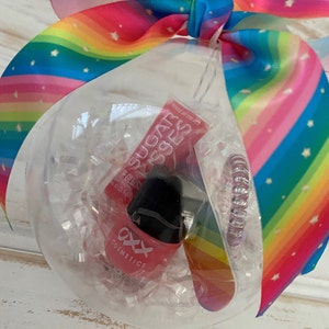 Puede incluir: Un adorno de pl&aacute;stico transparente relleno de papel de seda blanco y decorado con una cinta arco&iacute;ris. Dentro del adorno hay tres art&iacute;culos: un tubo de brillo de labios rosa etiquetado como "Sugar Kisses", una botella de esmalte de u&ntilde;as rosa etiquetada como "axx cosmetics" y una liga para el cabello de color arco&iacute;ris.