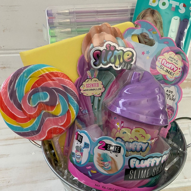 K&ouml;nnte beinhalten: Ein silberner Metalleimer gef&uuml;llt mit Schleim-Zubeh&ouml;r, darunter ein lila Schleimbeh&auml;lter, ein Lollipop, eine Packung duftenden Marshmallow-Schleims und eine Packung fluffigen Schleims. Die Verpackungen zeigen den Text "So Slime DIY", "Mach deinen eigenen fluffigen Schleim!" und "Fluffy Slime Shake".