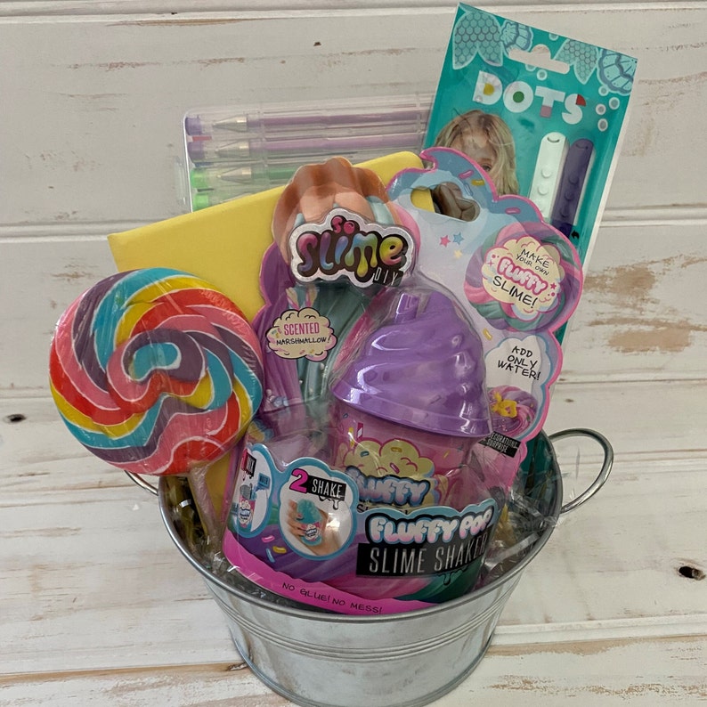K&ouml;nnte beinhalten: Ein silberner Metalleimer gef&uuml;llt mit verschiedenen Bastelartikeln, darunter ein Lollipop, Slime-Sets, Marker und ein Notizbuch. Die Slime-Sets haben eine farbenfrohe Verpackung mit den Worten "Fluffy Pop Slime Shaker" und "Mach dein eigenes fluffiges Slime!"
