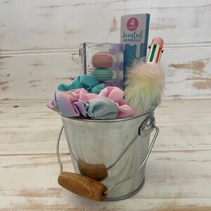 K&ouml;nnte beinhalten: Ein silberner Metalleimer gef&uuml;llt mit pastellfarbenen Haargummis, einer Packung duftender HB-Bleistifte, einem mehrfarbigen Stift und einem flauschigen Regenbogen-Pompon.