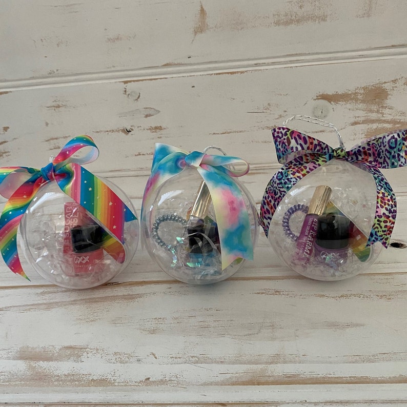 Puede incluir: Tres adornos de pl&aacute;stico transparente llenos de confeti de colores, purpurina y peque&ntilde;os regalos. Cada adorno est&aacute; decorado con una cinta diferente: arco&iacute;ris, tie-dye y estampado de leopardo.