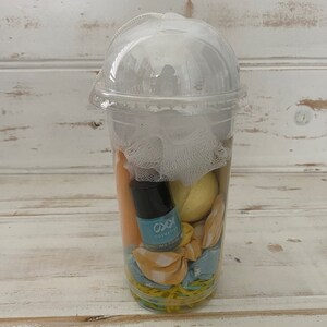 May include: A clear plastic cup filled with bath and beauty products. The cup contains a yellow bath bomb, a blue nail polish bottle with the text "OXX Cosmetics Nail Polish", a white mesh bath pouf, and a yellow and blue patterned fabric.