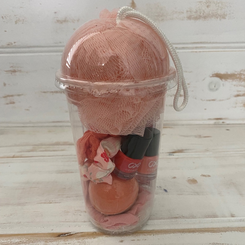 May include: A clear plastic cup filled with bath and beauty products. The cup contains a pink bath bomb, a pink bath puff, two bottles of nail polish, and a pink and white patterned hair scrunchie.