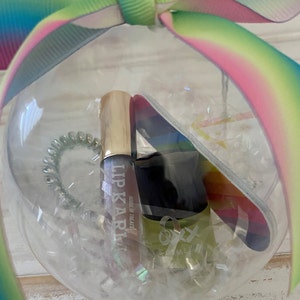 Puede incluir: Un adorno de vidrio transparente lleno de un tubo de brillo de labios, una lima de u&ntilde;as y una liga para el cabello. El tubo de brillo de labios es dorado y tiene el texto "Lip Karma" y "House of Treats". La lima de u&ntilde;as es de color arco&iacute;ris. La liga para el cabello es verde.