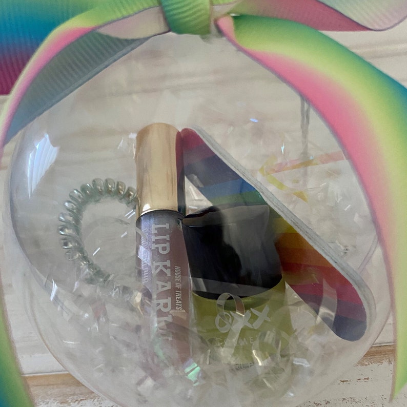 Puede incluir: Un adorno de vidrio transparente lleno de un tubo de brillo de labios, una lima de u&ntilde;as y una liga para el cabello. El tubo de brillo de labios es dorado y tiene el texto "Lip Karma" y "House of Treats". La lima de u&ntilde;as es de color arco&iacute;ris. La liga para el cabello es verde.