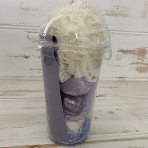 May include: A clear plastic cup filled with bath products. The cup contains a white bath puff, a purple bath bomb, a small bottle of bath oil, and a purple bath fizzy. The cup is decorated with a purple and white design.