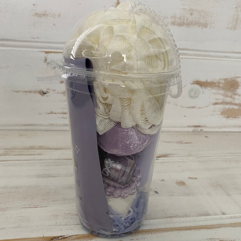 May include: A clear plastic cup filled with bath products. The cup contains a white bath puff, a purple bath bomb, a small bottle of bath oil, and a purple bath fizzy. The cup is decorated with a purple and white design.