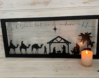 May include: Black framed Christmas decoration with a silhouette of the nativity scene, including the three wise men on camels, a star, and the text "O Come let us adore Him." A lit candle is in the foreground.