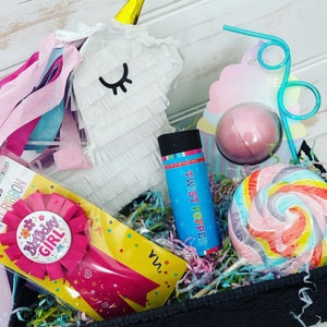 Pode incluir: Uma cesta de presente para uma festa de aniversário com uma piñata de unicórnio, uma pirulito rosa e azul, uma bomba de banho, um foguete de festa e uma fita de aniversário para uma menina.