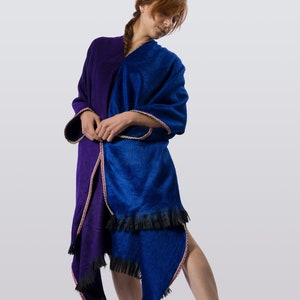 以下が含まれることがあります： 青い紫色のショールを着用した女性。ショールは黒いフリンジが付いており、体の周りに巻かれています。ショールには装飾的な縁取りがあります。
