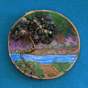May include: A handmade tree sculpture on a wooden disc. The tree has a wire trunk and branches, decorated with green, purple, and pink gemstones. The background shows a painted landscape with a river and path.