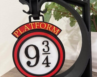 May include: Black 3D-printed sign featuring the words "PLATFORM" in gold lettering on a red banner. Below, a circular sign displays "9 3/4" in black. The sign is supported by a black stand with the words "Harry Potter" at the base.