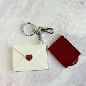 May include: A white envelope-shaped keychain with a red heart and a red square container. The keychain has a silver ring and clasp. The items are on a light pink and white marbled surface.