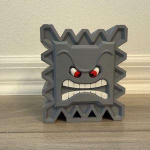 May include: A gray, 3D-printed Thwomp character from the Super Mario video game series. The block-shaped figure has a menacing expression with red eyes, a wide mouth, and jagged edges. The figure is displayed on a wooden surface.