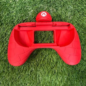 May include: Bright red video game controller grip with a brick pattern on the handles. The grip has a small red dome with a white "M" logo on top. The controller is designed to fit a handheld gaming device.