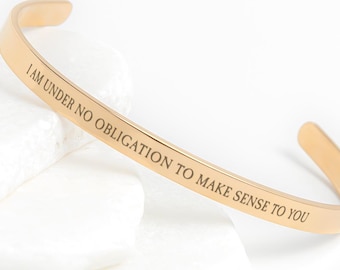 May include: A gold-colored cuff bracelet with the engraved text: "I AM UNDER NO OBLIGATION TO MAKE SENSE TO YOU." The bracelet is resting on white, smooth stones against a white background. This minimalist jewelry piece is a stylish accessory.