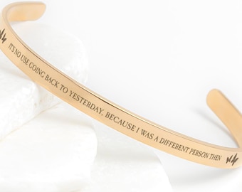 May include: A gold-colored cuff bracelet with the engraved text "IT'S NO USE GOING BACK TO YESTERDAY, BECAUSE I WAS A DIFFERENT PERSON THEN." The bracelet has a simple, elegant design and is displayed on white stones.
