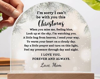 May include: A clear acrylic Christmas-themed light with a wooden base. The light has a semi-circular shape and features the text "I'm sorry I can't be with you this Christmas." The light is held by a hand and is surrounded by greenery.