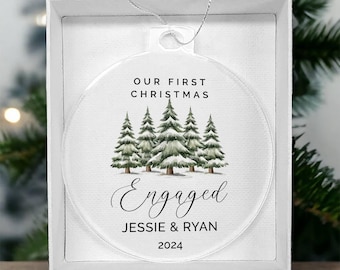 Puede incluir: Adorno navideño redondo y transparente con el texto "OUR FIRST CHRISTMAS" y "Engaged" con nombres y el año 2024. El adorno presenta una ilustración de acuarela de árboles de hoja perenne y se presenta en una caja blanca.