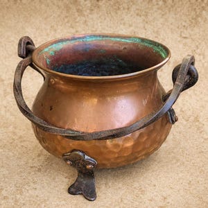 Puede incluir: Un caldero de cobre con textura martillada, con un asa oscura y retorcida y tres patas decorativas. El interior muestra una pátina azul y verde. Una pieza rústica y vintage.