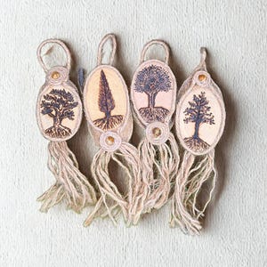 Peut inclure: Quatre porte-clés artisanaux sur le thème des arbres. Chaque porte-clés présente un pendentif ovale en bois avec un motif d'arbre dans des tons de marron et de bleu. Les pendentifs sont ornés de franges en jute et d'une petite perle dorée.