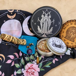 May include: A black cloth with moon phases and pink roses, a round black tin with a triple goddess design, a wooden divination board with French text, a small teal pouch, a dried herb bundle, and a metal tin containing herbal salt mix.