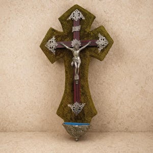 May include: A decorative wall cross with a silver crucifix figure. The cross is made of dark wood and is covered in olive green fabric. Silver filigree accents adorn the cross arms. A small, ornate silver bowl is at the base.