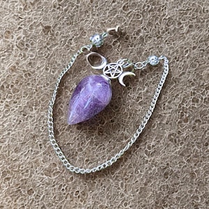 Può includere: Un pendolo di ametista viola con una catena d'argento e dettagli decorativi. Il pendolo presenta un cristallo a forma di goccia ed è collegato a una catena con un simbolo della triplice luna e una stella. La catena è attaccata a una chiusura.
