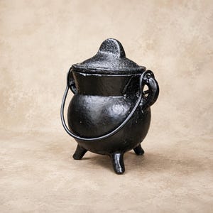 May include: A black cast iron cauldron with a textured surface, featuring three short legs, two side handles, and a matching lid. A thin, curved metal handle is attached to the sides, arching over the top of the round, deep cooking vessel.