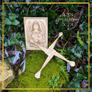 May include: A wooden St. Brigid's cross and a rectangular wooden plaque with an engraved image of a woman holding a torch and a lamb. The plaque has the word "BRIGID" on it. The items are on a bed of green moss and surrounded by green foliage.